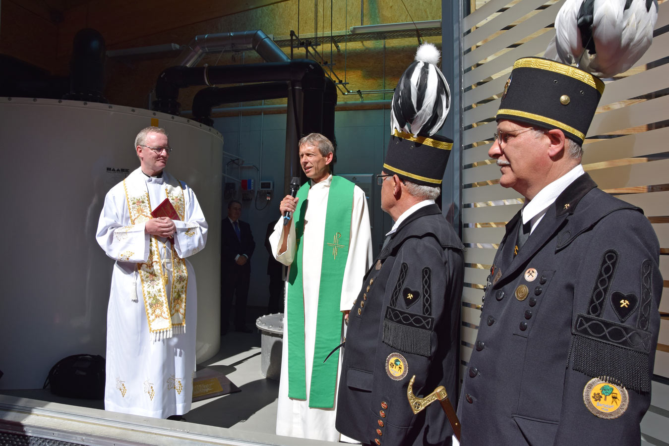 Ein Mann mit einem Mikrofon und einer Tscherpe steht in der Mitte des Bildes, ein Mann mit einer Bischofsuniform steht nehmen ihm genauso wie zwei Männer in traditionellen Uniformen